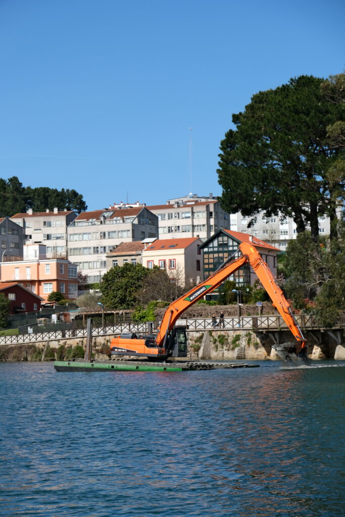 Natuurherstel en Waterbouw, Rental Referentie, Zicht: WK220 NG-5 werkzaam op water A Coruna, zijpontons aanwezig met spotpalen, zichtbaar oranje WK220 NG-5, achtergrond stad zichtbaar en zon op het water